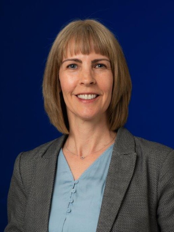Elizabeth stands infront of a dark blue background wearing a grey suit jacket and blue blouse. Her hair is straight at shoulder length, and she is smiling.