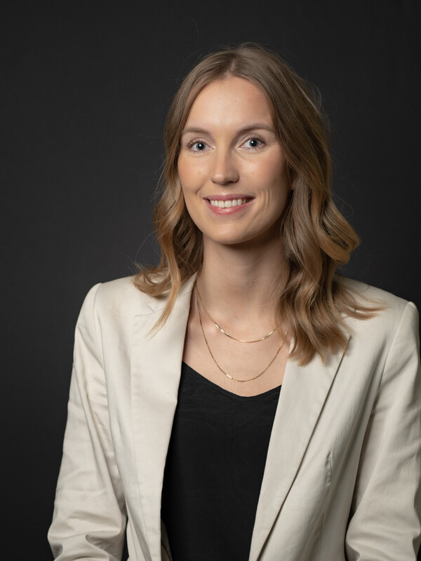 Victoria is standing against a black background, wearing a cream business jacket and black shirt; her hair is light brown, wavey, and down. She is smiling.